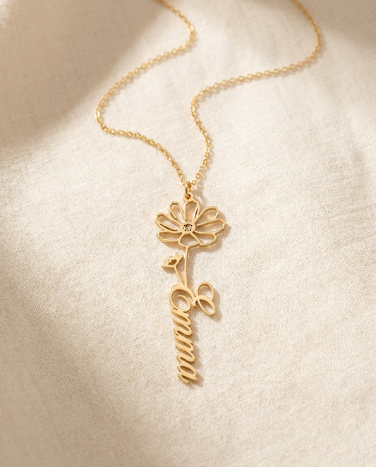 Gold-tone cut-out pendant shaped as an open-outline flower whose stem is the name Emma in flowing script, on a fine chain resting on cream linen