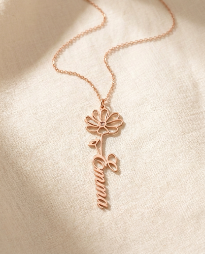Rose-tone birth-flower name pendant reading Emma in flowing script, on a fine chain resting on cream linen