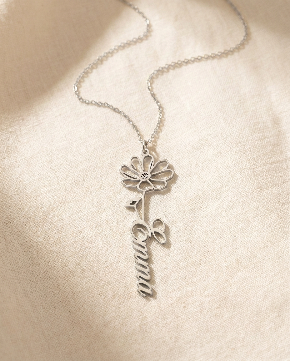 Silver-tone birth-flower name pendant reading Emma in flowing script, on a fine chain resting on cream linen