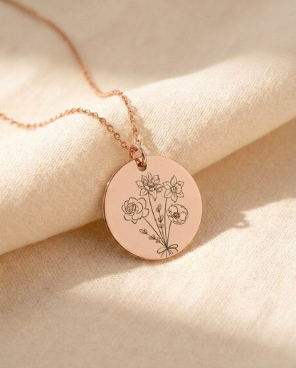 Rose-tone round disc pendant on a fine chain resting on cream linen, engraved with a bouquet of a rose, a poppy, a lily and a daffodil