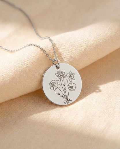 Silver-tone round disc pendant on a fine chain resting on cream linen, engraved with a bouquet of a rose, a poppy, a lily and a daffodil