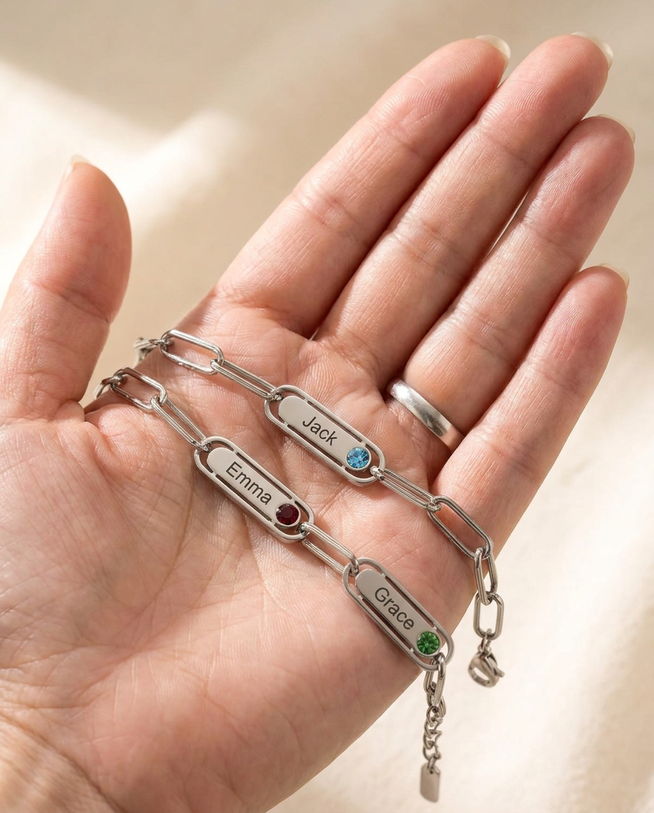 An open palm holding a silver-tone bracelet whose name bars are engraved Jack, Emma and Grace, each with a small coloured stone