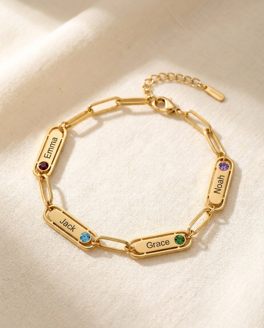 Gold-tone paperclip chain bracelet with four engraved name bars reading Emma, Jack, Grace and Noah, each with a small coloured stone, lying on cream linen in window light