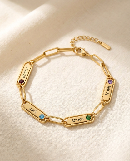 Gold-tone paperclip chain bracelet with four engraved name bars reading Emma, Jack, Grace and Noah, each with a small coloured stone, lying on cream linen in window light