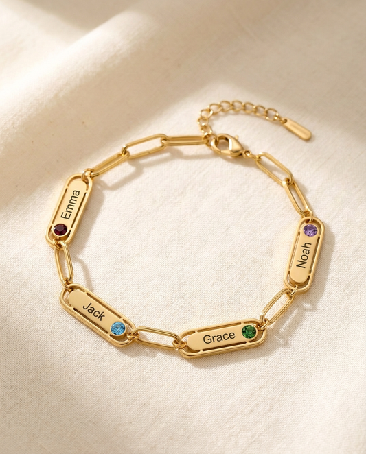 Gold-tone paperclip chain bracelet with four engraved name bars reading Emma, Jack, Grace and Noah, each with a small coloured stone, lying on cream linen in window light
