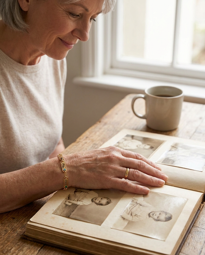 A grey-haired woman smiling over an old family photo album at a kitchen table, wearing a gold-tone bracelet with name bars engraved Jack and Grace