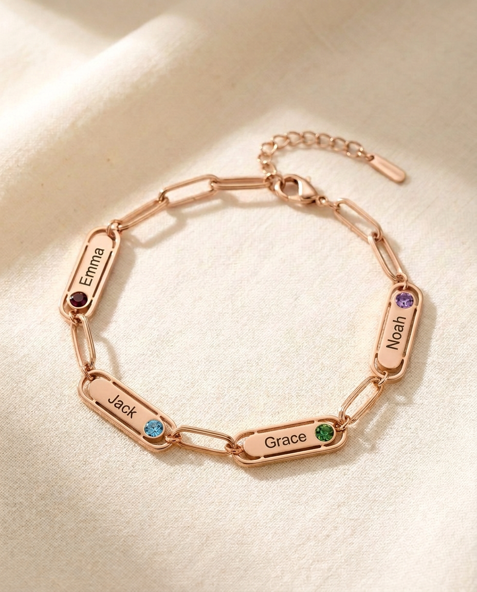 Rose-tone paperclip chain bracelet with four engraved name bars reading Emma, Jack, Grace and Noah, each with a small coloured stone, on cream linen
