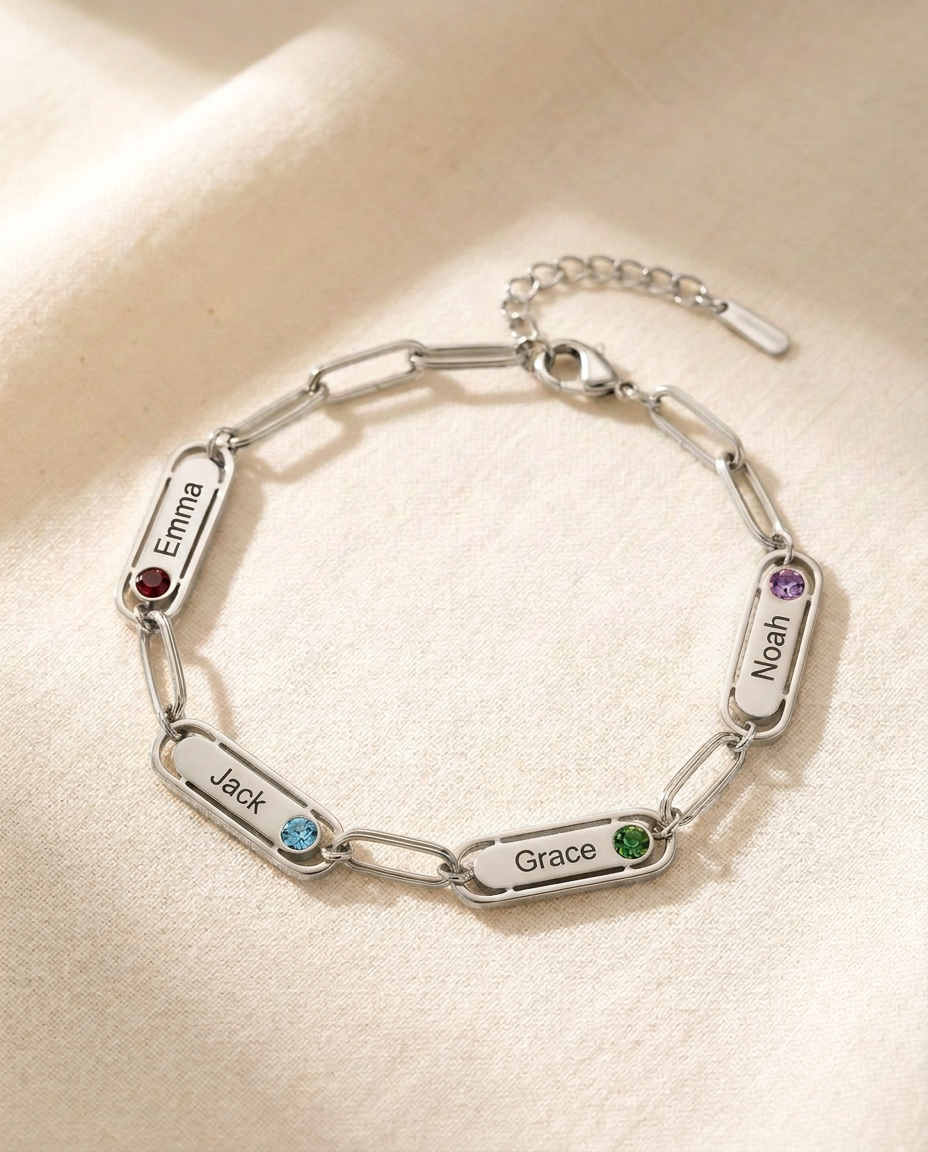 Silver-tone paperclip chain bracelet with four engraved name bars reading Emma, Jack, Grace and Noah, each with a small coloured stone, on cream linen