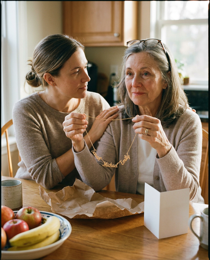 Mother at a kitchen table holding up a gold-tone necklace with the names Sarah, Michael and Emily, her adult daughter beside her with a hand on her shoulder, torn kraft paper and a blank card on the table