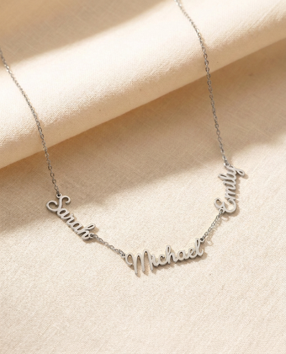 Silver-tone necklace with the names Sarah, Michael and Emily cut out in flowing script, linked side by side on a fine chain, lying on cream linen