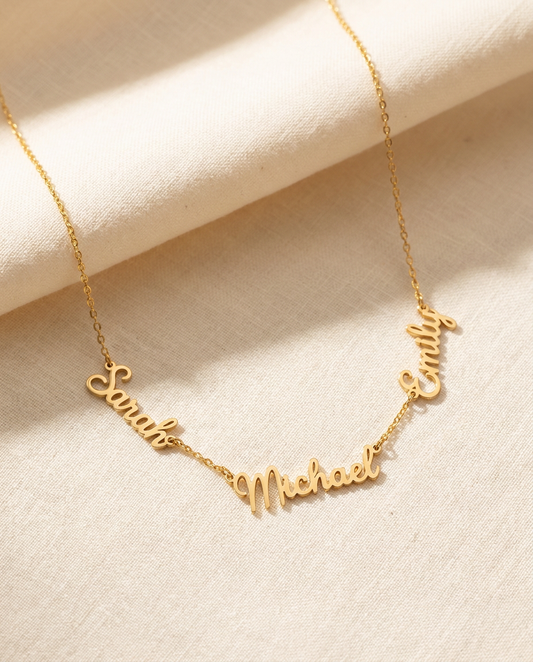 Gold-tone necklace with the names Sarah, Michael and Emily cut out in flowing script, linked side by side on a fine chain, lying on cream linen