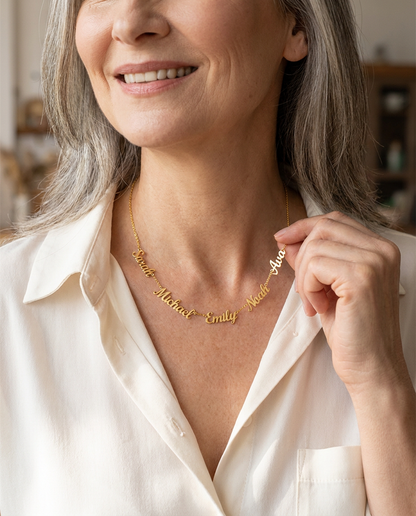 Smiling woman in her late fifties with silver-streaked hair and an ivory open-collar blouse, wearing a gold-tone necklace of five script names, Sarah, Michael, Emily, Noah and Ava, at her collarbone
