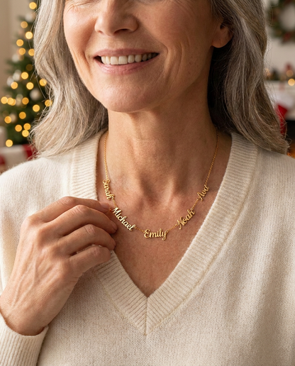 Smiling woman in a cream knit V-neck sweater wearing a gold-tone necklace of five script names, Sarah, Michael, Emily, Noah and Ava, with Christmas tree lights glowing behind her