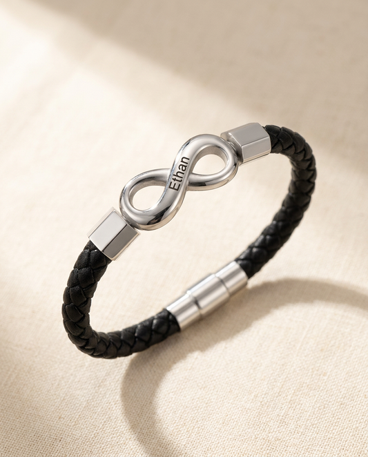 Braided cord bracelet with a steel-tone infinity link engraved Ethan, lying in an open curve on cream linen in soft window light