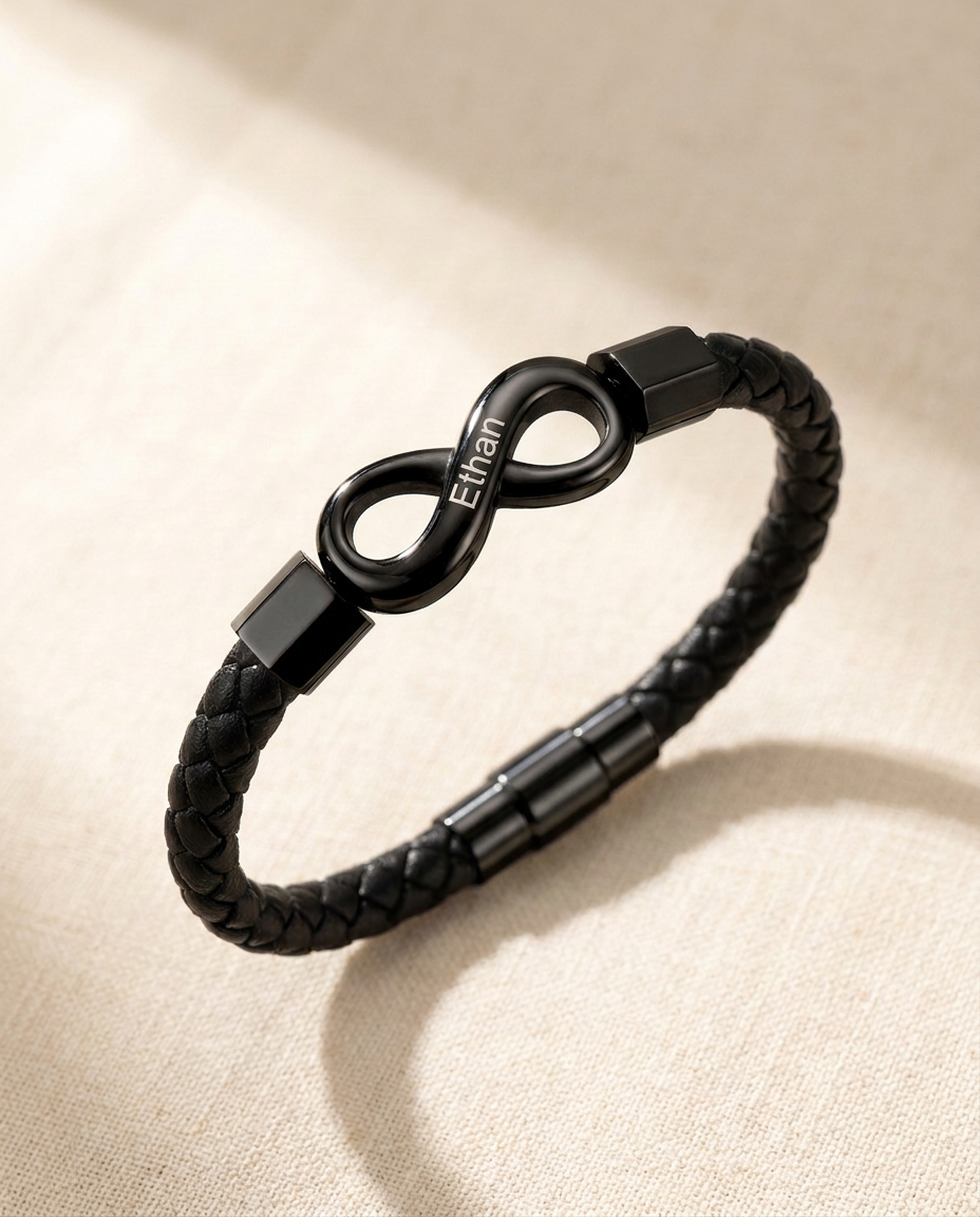 Braided cord bracelet with a black infinity link engraved Ethan, lying on cream linen in soft window light