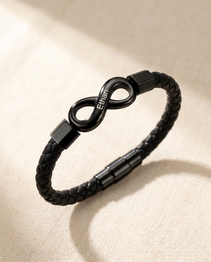 Braided cord bracelet with a black infinity link engraved Ethan, lying on cream linen in soft window light